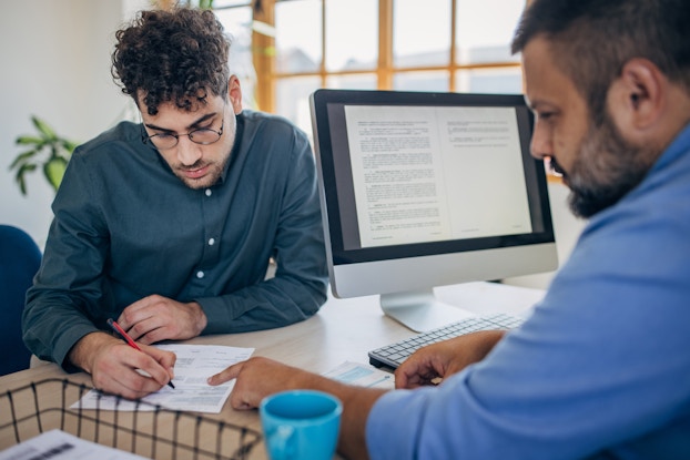  An angled shot of two men sitting on opposite sides of a desk in an office. The man on the left, seen from over the shoulder of the man on the right, is signing a piece of paper.  The man on the right is pointing at where the other man should sign. A computer monitor is sitting on the desk and facing the man on the right; its screen shows a two-page spread of a letter or contract.