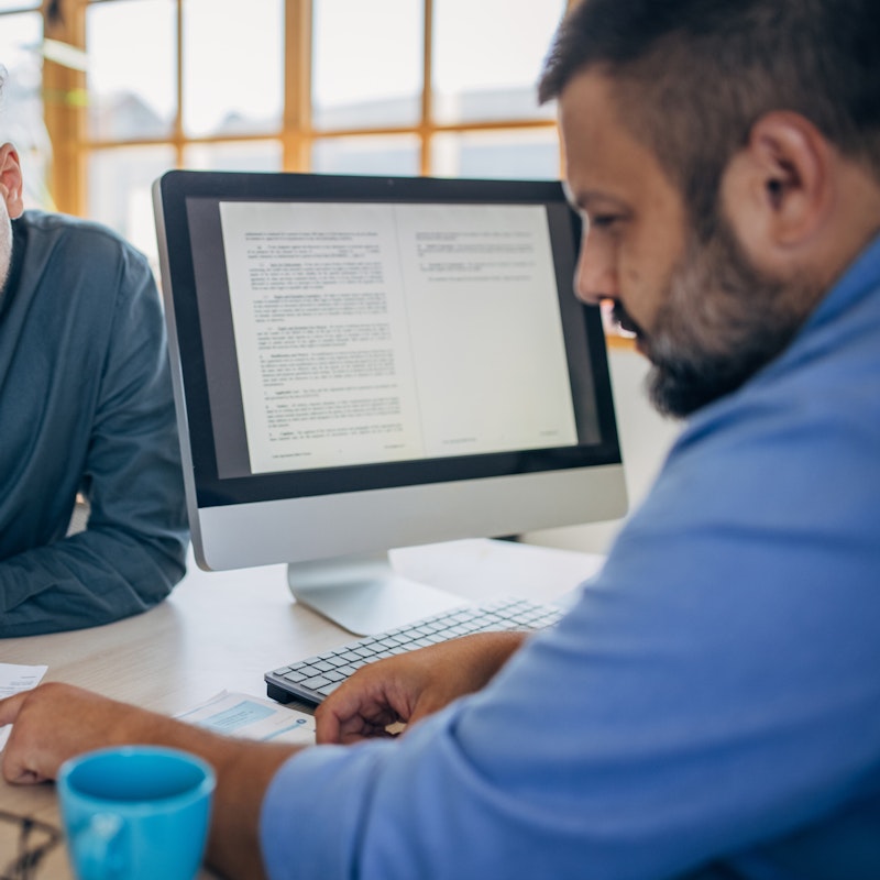 An angled shot of two men sitting on opposite sides of a desk in an office. The man on the left, seen from over the shoulder of the man on the right, is signing a piece of paper.  The man on the right is pointing at where the other man should sign. A computer monitor is sitting on the desk and facing the man on the right; its screen shows a two-page spread of a letter or contract.