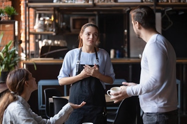  A young woman in an apron is berated by two angry customers. One of the customers is sitting and gesturing with one hand; the other customer, a man with a beard, is standing and holding an empty coffee cup. The waitress in the apron is centered in the picture. She looks at the standing man with a worried grimace on her face. 