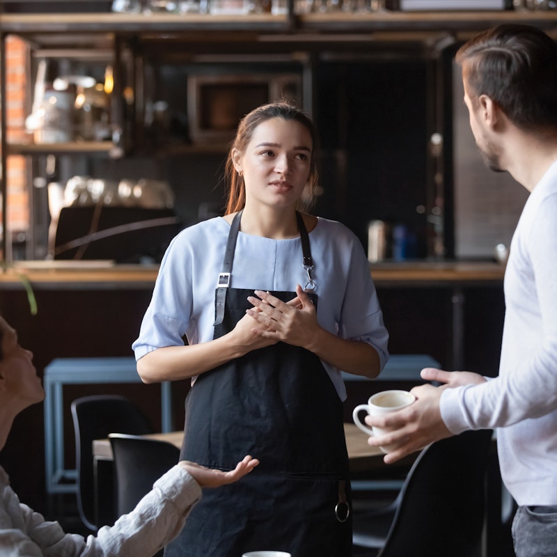  A young woman in an apron is berated by two angry customers. One of the customers is sitting and gesturing with one hand; the other customer, a man with a beard, is standing and holding an empty coffee cup. The waitress in the apron is centered in the picture. She looks at the standing man with a worried grimace on her face. 