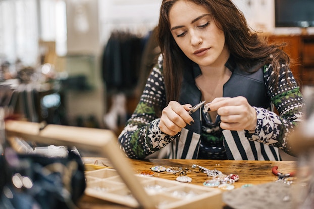  A woman sits at a table and uses a set of pliers to adjust a small piece of jewelry. Other pieces of jewelry -- namely pendants with various designs -- lay on the table before her, along with an open wooden box with several small compartments.