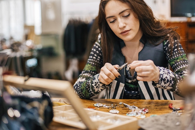 A woman sits at a table and uses a set of pliers to adjust a small piece of jewelry. Other pieces of jewelry -- namely pendants with various designs -- lay on the table before her, along with an open wooden box with several small compartments.