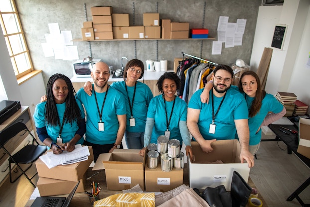 A diverse group of six people stands in a row behind a table piled with boxes of donations marked "food" and "clothes." The group members all wear turquoise t-shirts and white cards on black lanyards. The room around them is plain and industrial and filled with racks of clothing, stacks of books, more boxes and assorted office furniture.