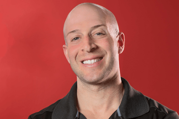 A head shot of Josh York, a smiling bald white man with gray eyes. He has his arms folded and is wearing a black polo shirt with the GYMGUYZ logo ("GYMGUYZ" in all-caps red-and-white font and "#1 in Home Personal Training" in smaller white font underneath) on the right side of the shirt.