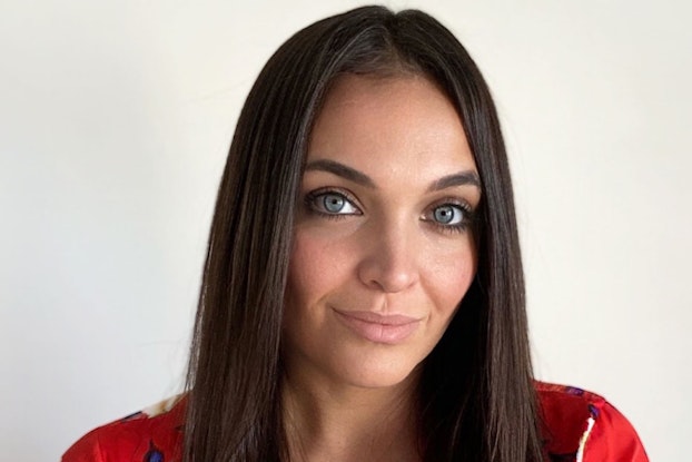 A medium shot of Kate Assaraf, a woman with shoulder-length brown hair and light blue-green eyes. She is shot from the waist-up and she wears a red blouse patterned with white flowers and dark blue leaves.