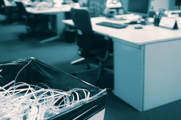  An office with an open floor plan. In the background, out of focus, are rows of desks holding computer monitors and keyboards. In the foreground, in focus, is a trash can filled with shredded paper.