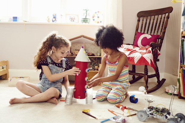 Two young girls work together to make a rocket ship made out of arts and crafts items. The rocket has a red body with a pointed white cap on top and three dials on the side. A few stickers shaped like flowers, hearts, and stars are stuck to the side and top of the rocket. The rocket's boosters on the bottom look as if they're made from three plastic soda bottles with the tops cut off. Behind the girls are a dollhouse and a rocking chair holding a red-and-white blanket and a red pillow bearing the letter C in white. On the floor next to the girl are scissors, colored pencils, a piece of paper, a roll of tape, a glue stick, and a small wheeled machine that resembles a lunar rover.
