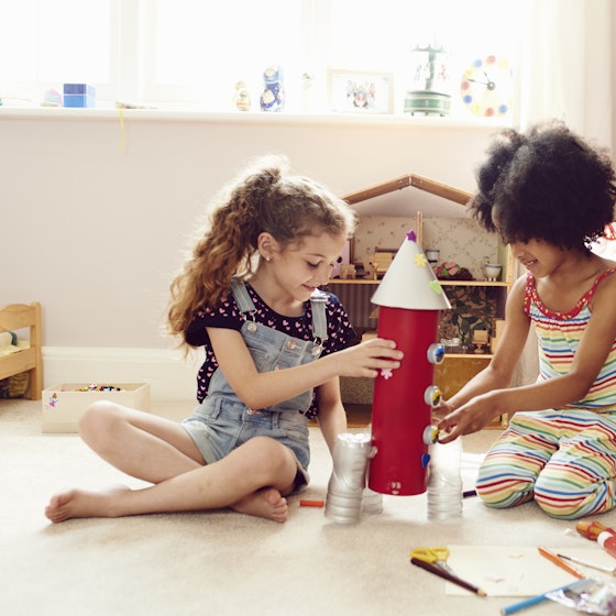 Two young girls work together to make a rocket ship made out of arts and crafts items. The rocket has a red body with a pointed white cap on top and three dials on the side. A few stickers shaped like flowers, hearts, and stars are stuck to the side and top of the rocket. The rocket's boosters on the bottom look as if they're made from three plastic soda bottles with the tops cut off. Behind the girls are a dollhouse and a rocking chair holding a red-and-white blanket and a red pillow bearing the letter C in white. On the floor next to the girl are scissors, colored pencils, a piece of paper, a roll of tape, a glue stick, and a small wheeled machine that resembles a lunar rover.