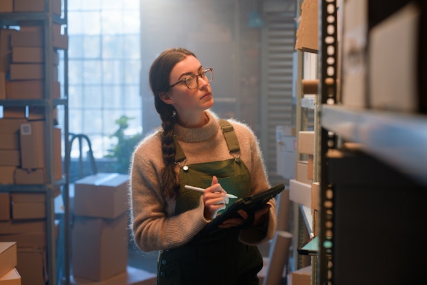 A young woman walks beween shelves in a small warehouse while making notes on a clipboard. The shelves are packed with cardboard boxes. The woman has long brown hair tied in a braid, and she wears glasses, a light brown turtleneck sweater, and olive green overalls.
