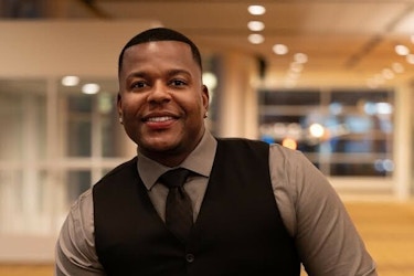 A shot of Daniel Johnson, owner of Levels, LLC. Daniel Johnson is a young Black man with short, dark hair. He's smiling and leaning back on a patterned yellow seat. He is wearing a gray collared shirt, black tie, black vest, and black trousers. 