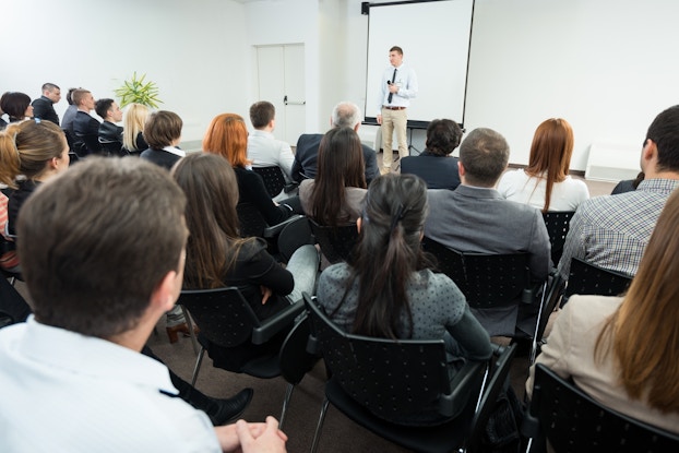 A wide shot of a large white room filled with about twenty people sitting in black chairs. The chairs and their occupants are facing away from the viewer and toward the front of the room, where a man in a white button-up shirt, black tie, and khaki pants is holding a microphone and standing in front of a projector screen.