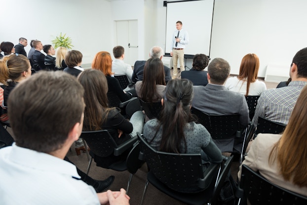  A wide shot of a large white room filled with about twenty people sitting in black chairs. The chairs and their occupants are facing away from the viewer and toward the front of the room, where a man in a white button-up shirt, black tie, and khaki pants is holding a microphone and standing in front of a projector screen.