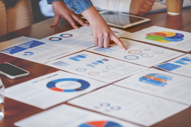 A polished wooden table covered with neatly laid out papers. The papers show various charts with sections and bars colored in shades of blue and orange. A hand attached to someone out of frame points to one of the charts (an orange circle showing a percentage).