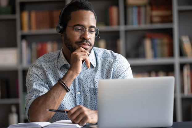 A man wearing glasses and headphones considers the contents of his laptop screen with one hand on his chin. In his other hand is a pen, held above an open notebook.