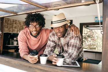  Two men lean on the ledge of the window of a food truck. One man, wearing glasses and a coral sweater, has his army around the shoulders of the other man, who wears a plaid shirt and a fedora. In front of them on the window ledge are white coffee cups with black lids and an electronic tablet. 