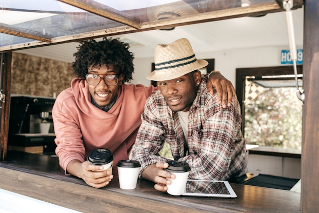  Two men lean on the ledge of the window of a food truck. One man, wearing glasses and a coral sweater, has his army around the shoulders of the other man, who wears a plaid shirt and a fedora. In front of them on the window ledge are white coffee cups with black lids and an electronic tablet.