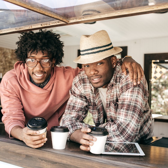 Two men lean on the ledge of the window of a food truck. One man, wearing glasses and a coral sweater, has his army around the shoulders of the other man, who wears a plaid shirt and a fedora. In front of them on the window ledge are white coffee cups with black lids and an electronic tablet.