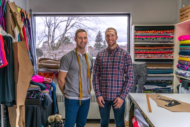  Two men stand smiling in a fabric store among shelves and racks of various brightly colored swatches of cloth. The man on the left stands with his hands behind his back and wears jeans and a gray-and-white polo shirt. He has bright yellow measuring tape draped around his neck. The man on the right wears jeans and a long-sleeved flannel shirt. Near him in the foreground is a table with a cut-out piece of khaki fabric, a yardstick and other measuring tools on top.