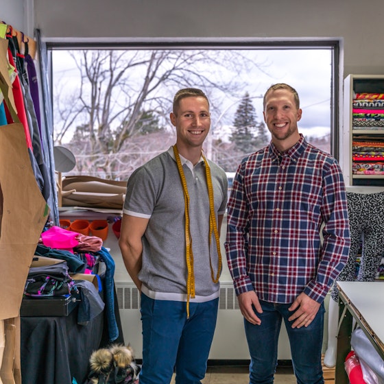  Two men stand smiling in a fabric store among shelves and racks of various brightly colored swatches of cloth. The man on the left stands with his hands behind his back and wears jeans and a gray-and-white polo shirt. He has bright yellow measuring tape draped around his neck. The man on the right wears jeans and a long-sleeved flannel shirt. Near him in the foreground is a table with a cut-out piece of khaki fabric, a yardstick and other measuring tools on top. 
