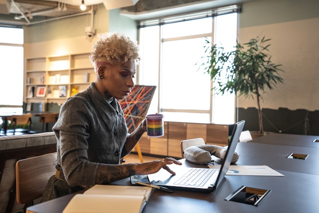 A tattooed woman sits at a desk and uses one hand to maneuver the trackpad on an open laptop. In her other hand, she holds a mug with a rainbow pattern on it. The woman has blonde curly hair cut short and shaved on the sides. She has a sleeve of tattoos on her right forearm and a word in cursive tattooed above one eyebrow. She wears a dark gray button-up shirt. The room behind her is a large office space with cream walls and a large window flanked by a potted tree and an abstract painting on an easel.