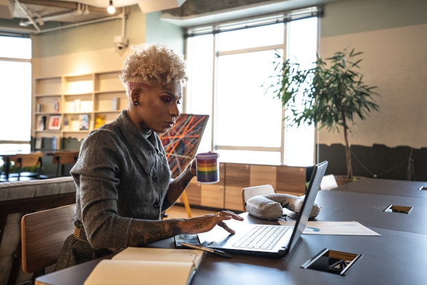  A tattooed woman sits at a desk and uses one hand to maneuver the trackpad on an open laptop. In her other hand, she holds a mug with a rainbow pattern on it. The woman has blonde curly hair cut short and shaved on the sides. She has a sleeve of tattoos on her right forearm and a word in cursive tattooed above one eyebrow. She wears a dark gray button-up shirt. The room behind her is a large office space with cream walls and a large window flanked by a potted tree and an abstract painting on an easel.