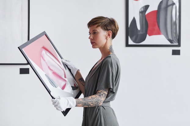  A woman wearing white gloves stands in a gallery holding a framed painting, looking thoughtful. The woman wears a gray dress and has short hair and a sleeve tattoo on her left arm. She examines an abstract painting made of pink, white and gray slashes and strokes.