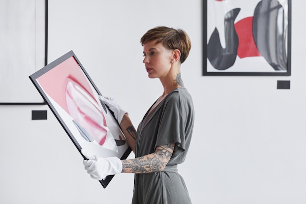 A woman wearing white gloves stands in a gallery holding a framed painting, looking thoughtful. The woman wears a gray dress and has short hair and a sleeve tattoo on her left arm. She examines an abstract painting made of pink, white and gray slashes and strokes.