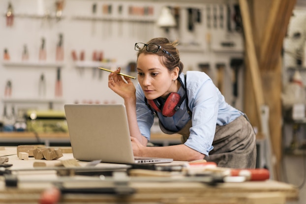  A woman in a carpenter's workshop leans over a wooden counter to look at something on the screen of an open laptop. The woman has brown hair pulled back on her head and she holds a pencil in one hand. She wears red safety headphones around her neck, glasses on top of her head, and a light blue button-up shirt under a weathered gray apron. The wooden counter is cluttered with pieces of wood in various shapes and sizes.