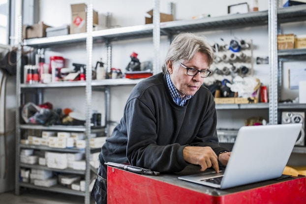 A man leans on a counter and looks at an open laptop while using one finger to work the keyboard's track pad. The man is older, with gray hair, and he is wearing a dark gray sweater over a blue-and-white checkered shirt. Behind him, out of focus, are a few steel shelving units holding automotive and mechanical equipment, some small white boxes, and red-and-black cans.