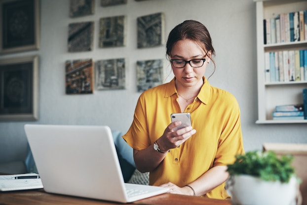A woman looks at her smartphone. On the desk in front of her is an open laptop.