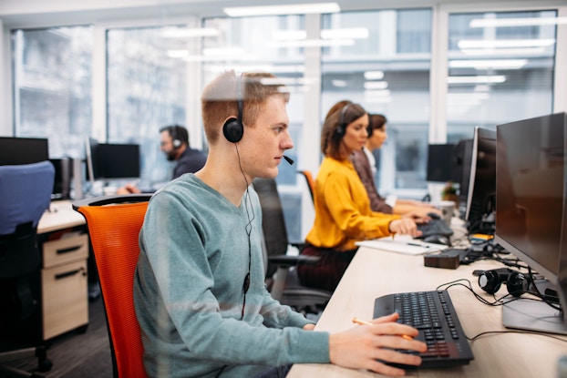A group of call center workers are pictured sitting at their desks. There is a young man at the forefront who is wearing a headset, looking at his computer screen, and typing on the keyboard.