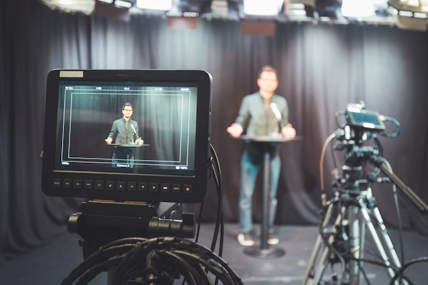  An electronic tablet on a tripod films a man standing at a podium and speaking into a microphone. The man, who wears a muted green zipped jacket and jeans, can be seen on the screen of the tablet and out of focus in the background. He stands in front of black curtains. Another tablet on a tripod can be seen off to one side, slightly out of focus.