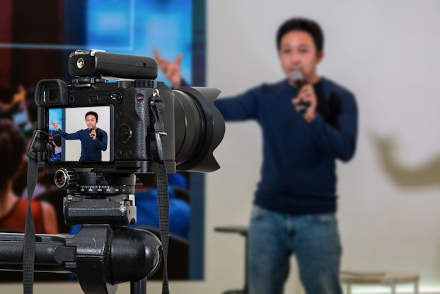  A camera, in focus, recoreds a man speaking on a microphone and gesticulating in front of a blank wall. The man can be seen on the camera's viewscreen and is shown out of focus in the background. To the left of the man is a screen showing the backs of members of an audience.