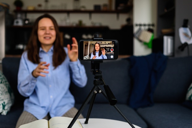 A smartphone positioned horizontally on a tripod films a woman speaking and gesturing. In the background, out of focus, is the woman herself.