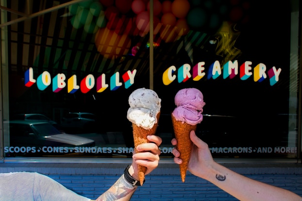  A short of two arms with hands holding ice cream cones in front of a Loblolly Creamery store window. The window has the words "LOBLOLLY CREAMERY" stenciled on it in white text with a multicolored 3D effect. The words "SCOOPS CONES SUNDAES SHAKES MACARONS AND MORE" are stenciled in smaller blue text along the bottom of the window. The hand on the left holds an ice cream cone with two scoops of a white ice cream with chocolate chips. The hand on the right has two scoops of a pink ice cream.