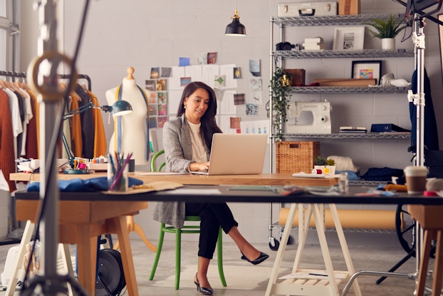 A woman sits at a long table in a large studio and works on a laptop. In the background are a grid of photos and papers pinned to the wall, a rack of clothing, a dress form used for pinning clothes, and a shelving unit holding a sewing machine, a few framed photos, and bundles of cloth.