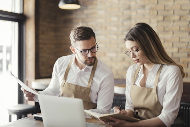 A man and a woman, both wearing glasses, white button-up shirts, and khaki aprons, sit side-by-side at a table. The woman sits on the right; she has long dark blonde hair and is writing something in a book with a tan cover. The man, who has a neatly trimmed beard and brown hair, is holding a slim stack of papers and looking over at what the woman is writing.