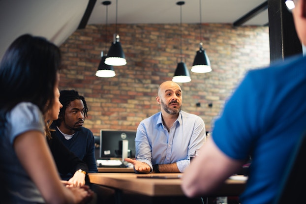 A business meeting is pictured. An older man, a manager, is speaking to employees. He is gesturing with his hands. The employees are looking at him and listening to him.