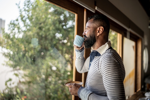 Person sipping coffee inside a home while gazing out the window into nature.