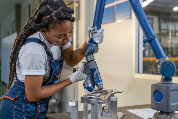 A woman on a manufacturing floor holds a robotic arm and aims it at a small sheet of glossy metal. The robotic arm is blue and has a needle-tipped nozzle at its end. The woman has long dark hair in thick braids. She wears blue overalls, a white T-shirt, and white gloves.