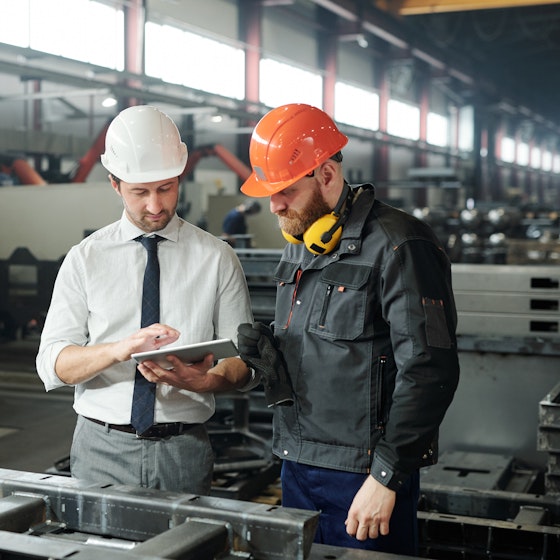  Two men wearing hard hats stand side by side in a warehouse filled with various machines and steel tables. Both men look at an electronic tablet held by the man on the left. 