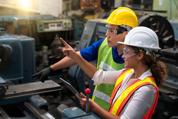  A woman and a man stand among industrial machinery. Both of them wear hard hats, protective goggles and neon high-visibility vests. The woman holds a clipboard and points off into the distances. The man wears black rubber gloves and points down at part of one of the machines. Near the woman's hand holding the clipboard is a lever with a bright red ball on top.