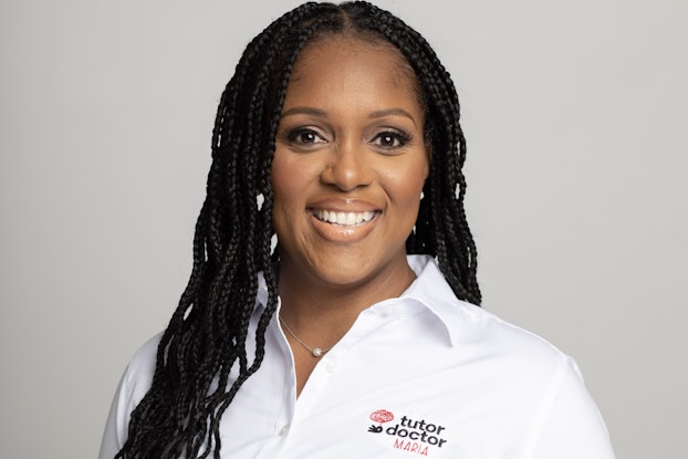 A headshot of Maria Washington, Co-owner of Tutor Doctor in Evans, Georgia. Maria is a smiling Black woman with long braided hair and dark brown eyes. She is wearing a white polo shirt with "tutor doctor" in black lower-case letters and "MARIA" in upper-case red letters stitched on the left side of the chest.