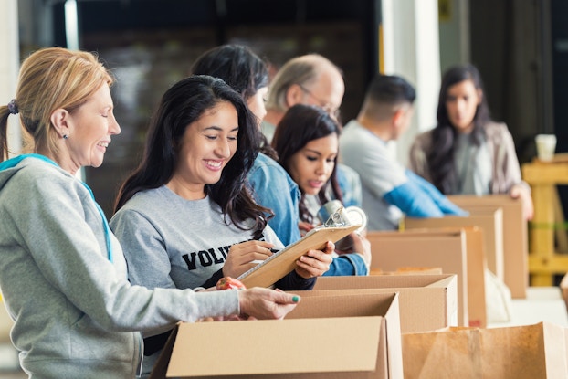 A line of people, each of them wearing a light gray hoodie or T-shirt, extends away from the viewer. Each person stands in front of an open cardboard box, which they are packing. Closest to the camera are two women. The one on the left has her hair in a ponytail and is laughing. The one on the right is writing something on a clipboard and smiling.