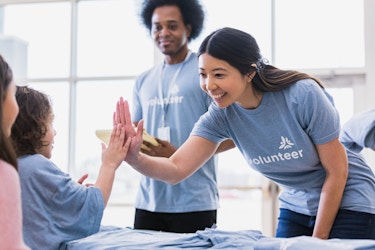  A young woman reaches across a table and high-fives a child. The woman and the child, as well as the young man standing behind the woman, are wearing light blue T-shirts. The T-shirts on the woman and the man say "volunteer" in white and have a symbol over the heart. The symbol is a triangle made up of several brushstrokes. The table is covered with folded light blue T-shirts. 