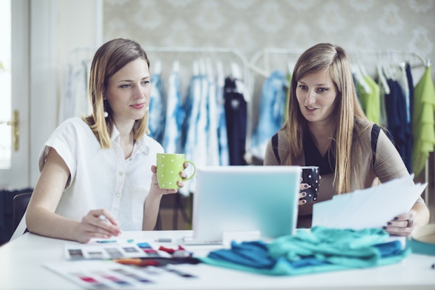 Two women holding coffee mugs sit at a table and look at the screen of an open laptop. The woman on the left has shoulder-length dark blonde hair; she holds a lime green mug covered with white polka dots and wears a short-sleeved white button-up shirt. The woman on the right has long dark blonde hair; she holds a black mug with white polka dots and wears a black blouse. The table also holds several papers and a few fabric swatches in various shades of blue. In the background are a few clothing racks filled with dresses on hangers.