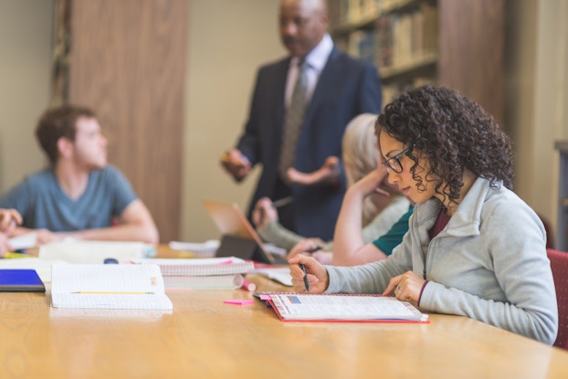  A woman wearing glasses reads from an open textbook. She's sitting at a table in a library. In the background are fellow students, also sitting with open books, and a lecturer in a suit, who is standing and talking.