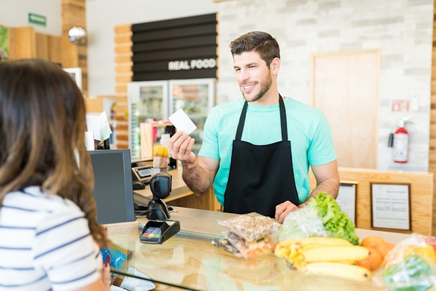 A man in an apron stands behind a checkout counter and holds up a white card. In the foreground, the customer, a woman with dark hair, stands on the other side of the checkout counter, facing away from the camera. On the checkout counter are various groceries, including a bunch of bananas, several oranges, and a bag of nuts.