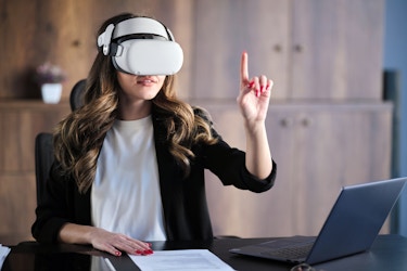  A woman wearing a VR headset sits at a desk and reaching up with one hand, a finger extended to poke or tap at something that only she can see. The table she sits at also holds an open laptop and a piece of paper covered with printed text. 