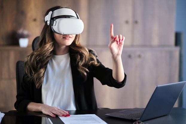  A woman wearing a VR headset sits at a desk and reaching up with one hand, a finger extended to poke or tap at something that only she can see. The table she sits at also holds an open laptop and a piece of paper covered with printed text.